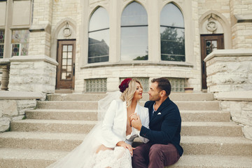 elegant and stylish bride with light curly hair dressed in a white dress, a white jacket and a red beret sitting on a stairs in a summer garden on the background of old big houses along with her