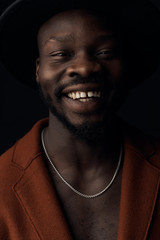 Dark key portrait of handsome smiling man with beard. Man dressed in brown coat and black hat, with silver neck chain. Studio shot, black background