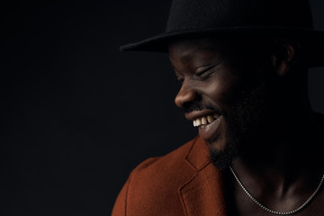 Dark key close-up portrait of handsome smiling man with beard in profile. Man dressed in brown coat and black hat, with silver neck chain. Studio shot, black