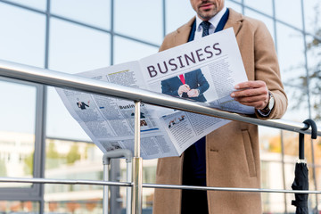 cropped view of businessman standing in beige coat and reading business newspaper