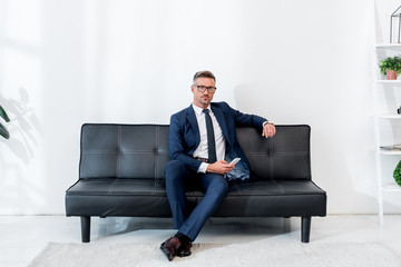 handsome businessman in suit sitting on sofa and holding smartphone