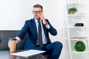 selective focus of handsome businessman in glasses talking on smartphone and holding paper cup