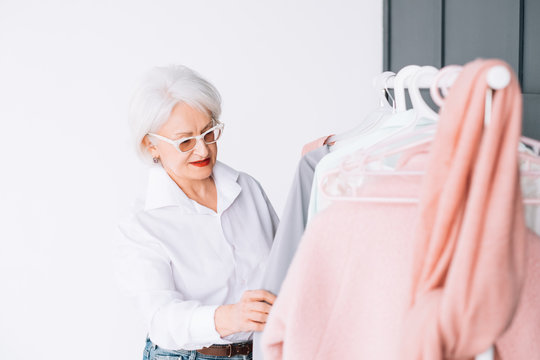 Senior Fashion. Stylish Showroom. Smart Elderly Lady In Glasses Looking Through Rack Of Clothing.