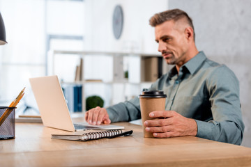 selective focus of paper cup in hand of businessman using laptop in office