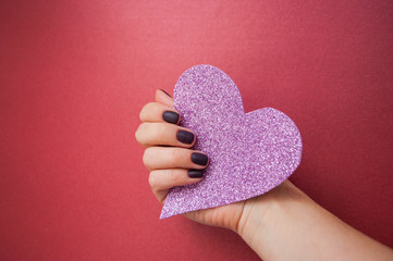 Young woman's hand with beautiful manicure on dark red background holding pink glittering heart.