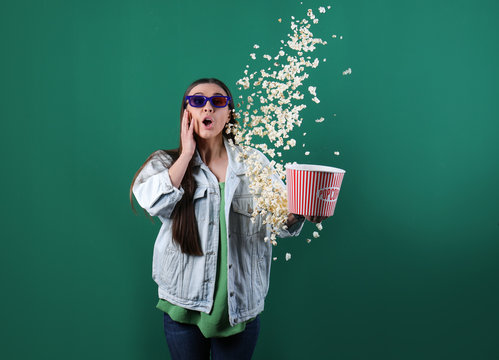 Emotional young woman with 3D glasses throwing popcorn on color background