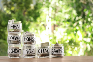 Glass jars with money for different needs on table against blurred background, space for text