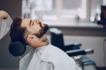 handsome young bearded guy sitting in an armchair in a beauty salon and the girl around him wipes his neck with a towel