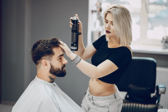 handsome young bearded guy sitting in an armchair in a beauty salon and the girl around him spray on his hair hairspray
