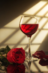 a glass of red wine and a red rose on the table with reflection, light from the window