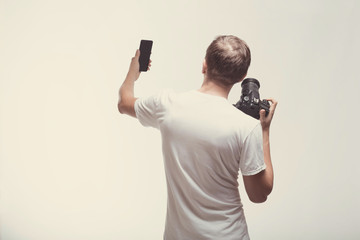 Emotional Man with camera isolated on light background. Young man hold digital camera and smartphone. Lifestyle, travel and technology concept. Standing back boy in white t-shirt making selfie