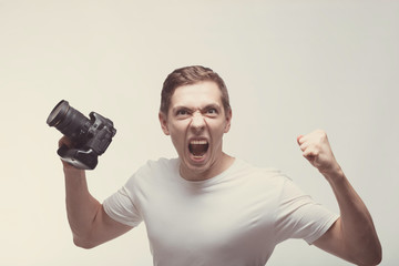 Angry emotional Man with camera isolated on light background. Young man holding digital camera and screaming. Lifestyle, travel and technology concept. Mad boy in white t-shirt with terrific face