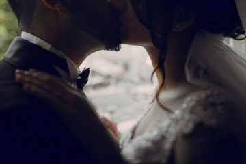 A beautiful young Turkish man wearing a black suit and a beard standing near windows along with his beautiful bride in a white dress 