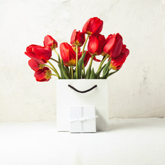White Gift Bag, small white gift box, petals and bouquet of red tulips on a light stone background. The concept of an offer for an engagement or a marriage