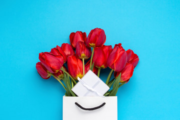 White Gift Bag, a small white gift box and a bouquet of red tulips on a blue background. Concept Offers an engagement or marriage, shopping