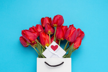 Bouquet of brightly red tulips, a small white gift box with a red heart in a white gift bag on a blue background. Conception of congratulations and a gift. Flat lay, top view
