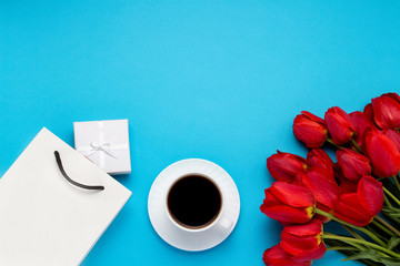 White Gift Bag, a small white gift box, a white cup with black coffee and a bouquet of red tulips on a blue background. Concept Offers an engagement or marriage, shopping