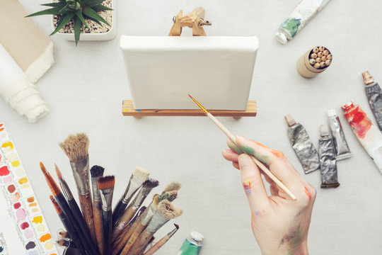 Painter holding a paintbrush in his hand. Canvas on the easel and artistic equipment on desk. Top view.
