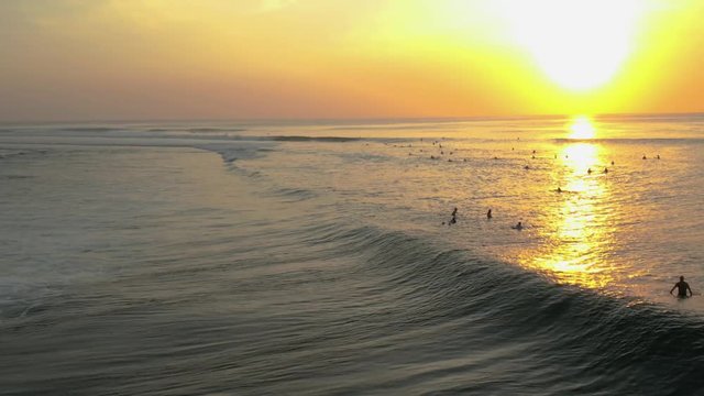 Moving aerial shot over Bali sea. Mellow waves crash into the shore. People swim and enjoy the tropical paradise as the sun sets in the background.