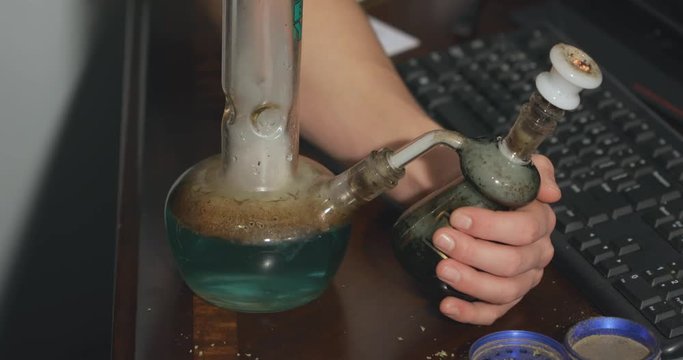 A hand holds and removes an ash catcher as a bowl of marijuana is smoked on an office desk.