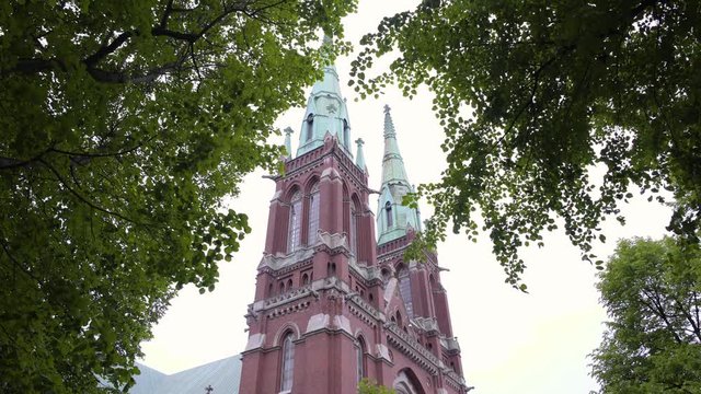 Static shot of St Johns Church, filmed under trees, green leaves and branches, waving in the wind, on a cloudy, summer day, in Ullanlinna, Helsinki, Finland