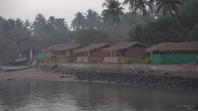 Panning shot across row of grass thatched colourful beach huts on the shoreline of Indian, Goa beach. View of coastal waters in foreground.