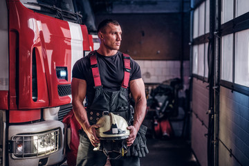 Muscular fireman holding a protective helmet in a garage of a fire department, standing next to a...