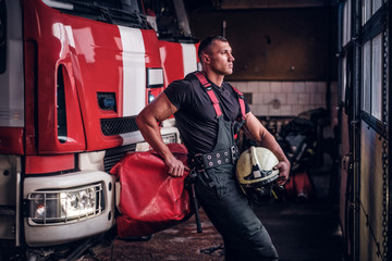 Muscular fireman holding a protective helmet in a garage of a fire department, leaning on a fire...