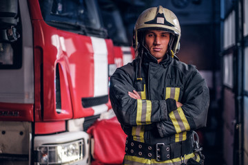 Confident fireman wearing protective uniform standing next to a fire engine in a garage of a fire...