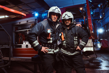 Two firemen hug and looking at the camera standing near the fire truck at night