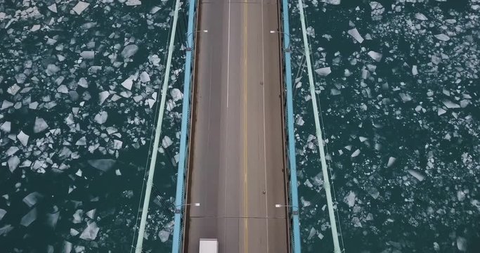 Transport trucks crossing the Ambassador Bridge to Detroit over icy blue water. Aerial pan up.