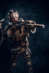 Elite unit, special forces soldier in camouflage uniform holding an assault rifle with a laser sight and aims at the target. Studio photo against a dark textured wall