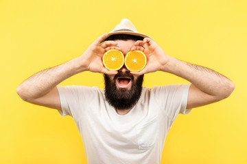 Studio portrait of a bearded man holding two halfs of orange citrus fruit in hands, covering his eyes, on yellow background, summer mood, happiness fun concept