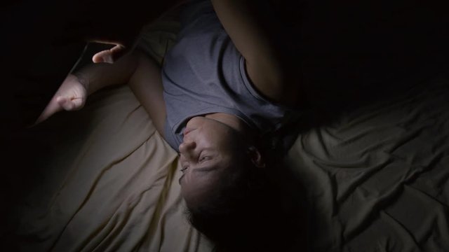 woman is lying in bed in night and viewing social nets news by tablet with wifi in home, light falling on face