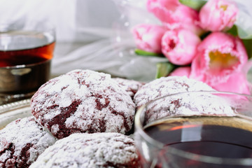 Red cracked cookies covered with white icing sugar on a silver old tray next to a cup of green tea in a glass cup with a silver cup holder