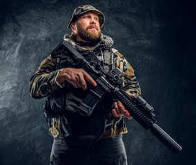 Brutal man in the military camouflaged uniform holding an assault rifle. Studio photo against a dark textured wall