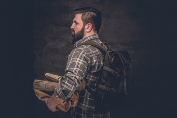 Portrait of a bearded woodcutter with a backpack dressed in a plaid shirt holding firewood. Studio photo against a dark textured wall