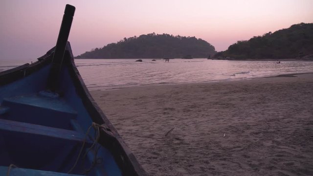 Goa, India, Palolem beach at sunset with pink violet sky and blue wood boat on the foreground. Waves moving in front of silhouette hills in distance.