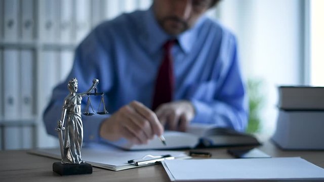 Lawyer writing notes on clipboard note paper in law office, selective focus on statue of Lady Justice