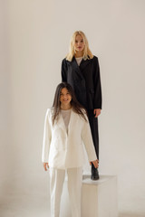 two women model posing for a photo. The blonde is dressed in a black suit and the brunette is dressed in a white suit. Stylish photo for advertising showroom or women's clothing store