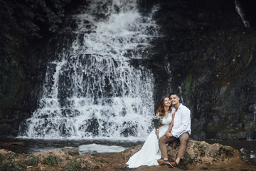 Fototapeta na wymiar Young couple in love bride and groom, wedding day near a mountain waterfall. Enjoy a moment of happiness and love. Bride in a luxurious wedding dress