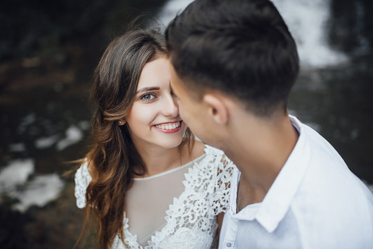 Young wedding couple of pretty girl or woman with hair in white bride dress and handsome man near beautiful grand waterfall in mountain