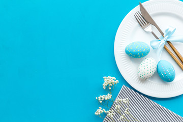 Easter table decoration in blue colors. Plate, cutlery, painted eggs and dry white flowers, tablecloth on blue background top view copy space