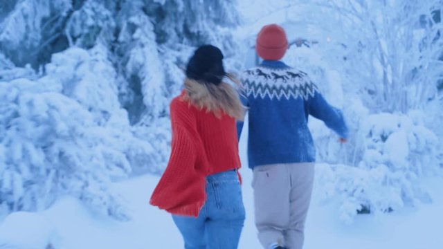Authentic couple of millennial hipsters, boyfriend and girlfriend hold hands and run through magical snow covered forest path towards bus, laugh and giggle on road trip getaway