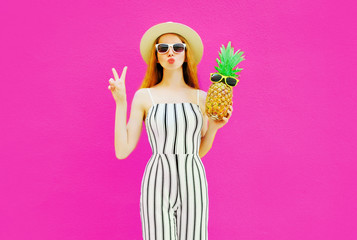 Portrait stylish woman sending sweet air kiss with pineapple in summer round straw hat, white striped jumpsuit on colorful pink wall background
