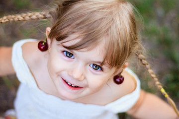 Portrait of cute little girl with green eyes playing with cherry in garden in sunny summer day