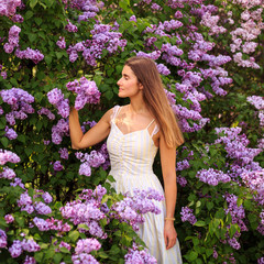 Beautiful young woman enjoying the smell of lilac in blossom garden in sunny spring day. Pretty female and flowers. Aromatherapy and springtime concept