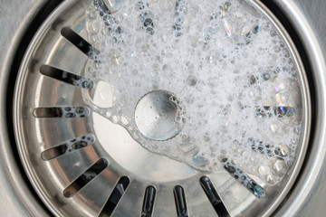 White foam with bubbles of cleane in a washbasin. Drain hole with soap bubbles in metal sink, macro view. Mechanically adjustable drain plug closeup.