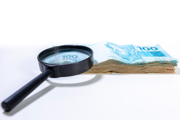 Stack of Brazilian Real banknotes with a magnifying glass resting on a corner and focussing on part...