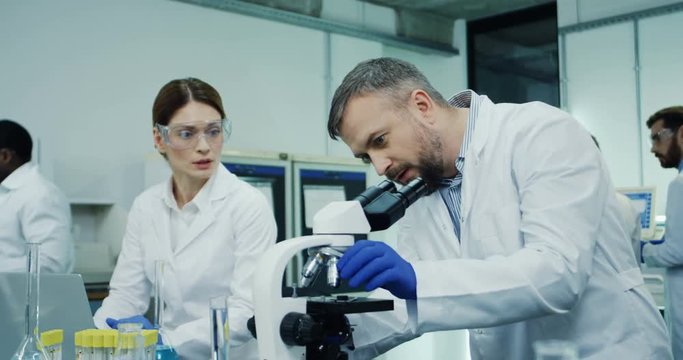 Male and female Caucasian colleagues working at the medical or pharmaceutical laboratory at the laptop computer and microscope while discussing their tests and results.